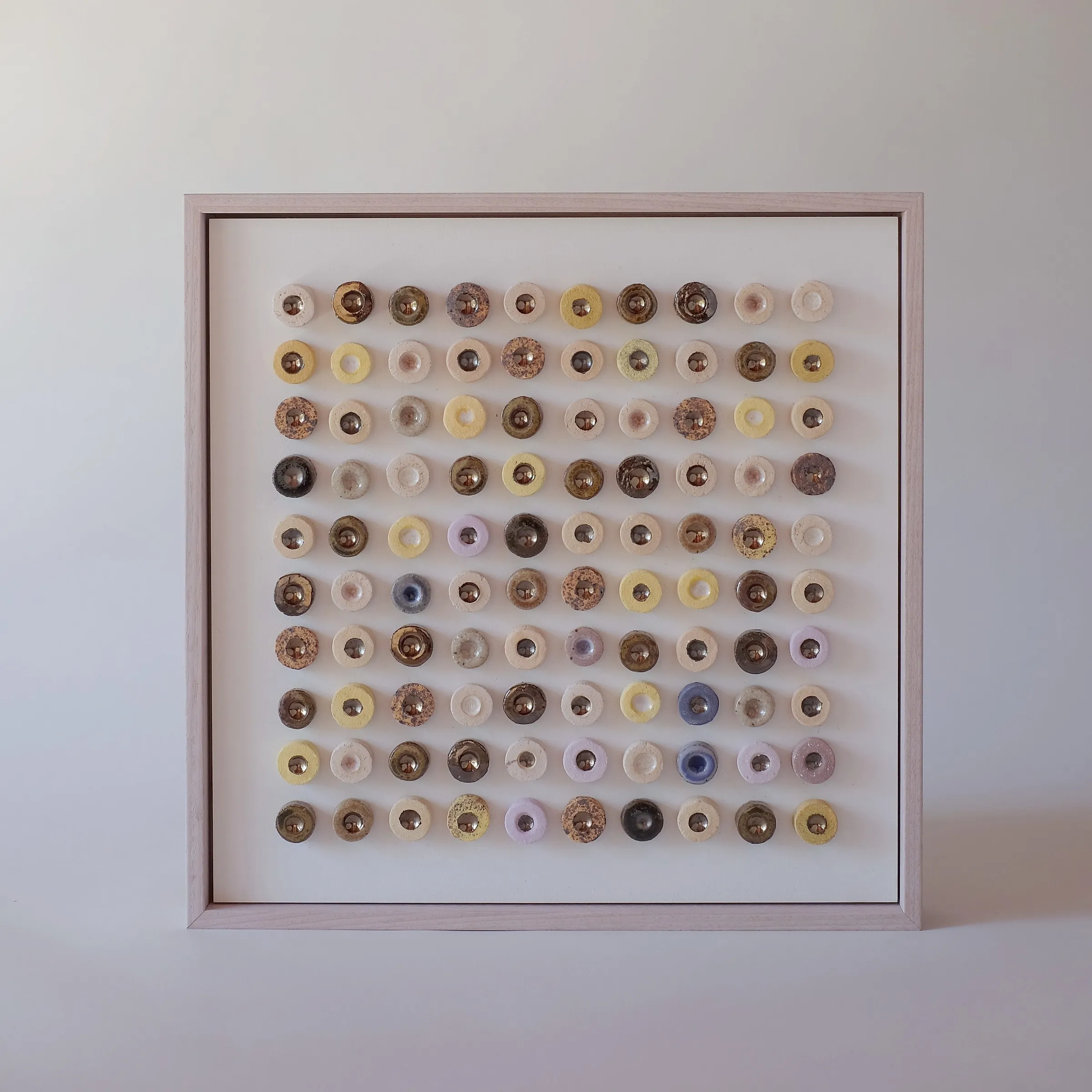 Dot Drift Duo (Suvarna) by Minaal Lawn, a framed grid of one hundred cream, gold and yellow glazed ceramic discs with lustre centres